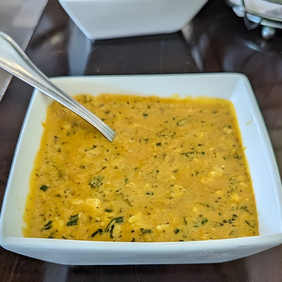 A white rectangular bowl filled with a yellow Indian curry, garnished with green herbs and a spoon.